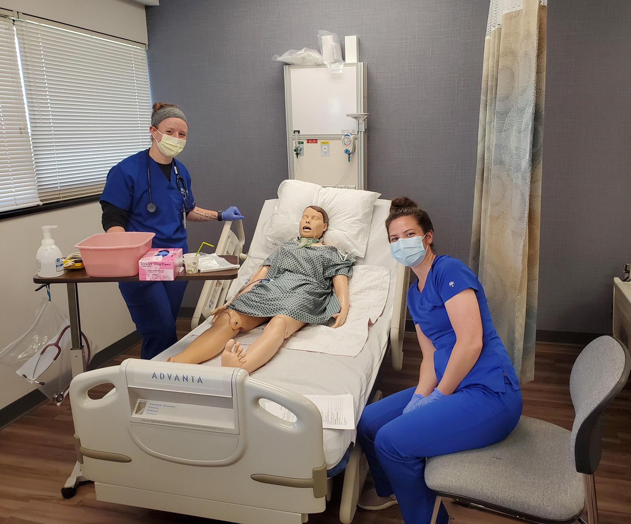 Two Herzing nursing students beside bed with simulation dummy