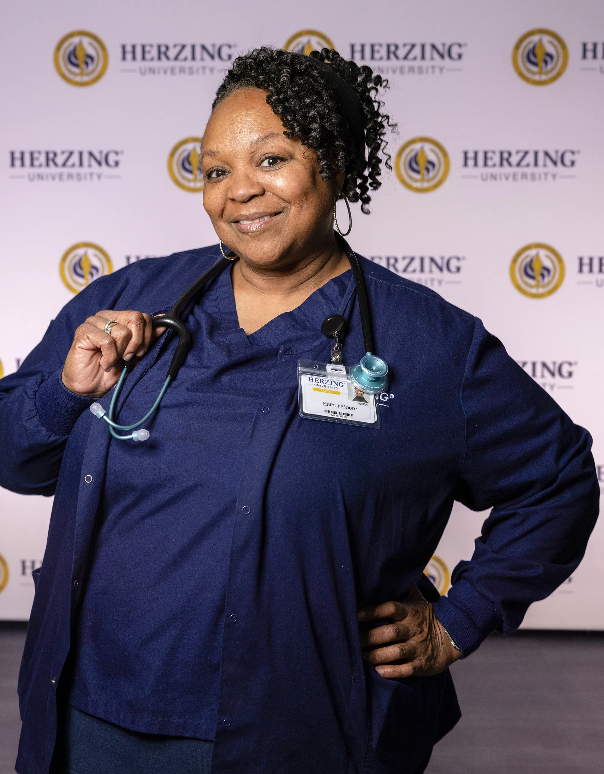Herzing University nursing student in navy scrubs wearing a stethoscope and smiling in front of a Herzing University backdrop.