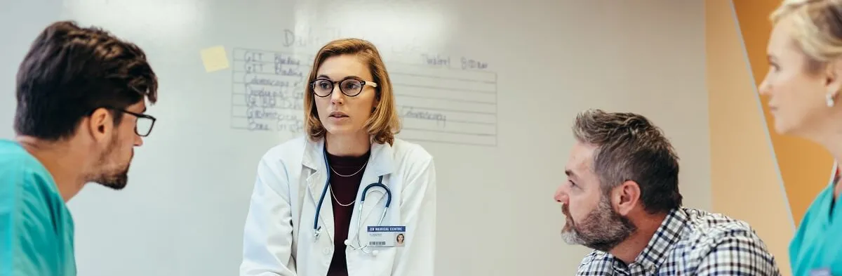 A female nurse administrator who went from associate degree RN to NLA wearing a white coat and stethoscope speaks to three colleagues during a team discussion. Two men and a woman listen attentively around a table, with a whiteboard containing medical notes visible in the background.