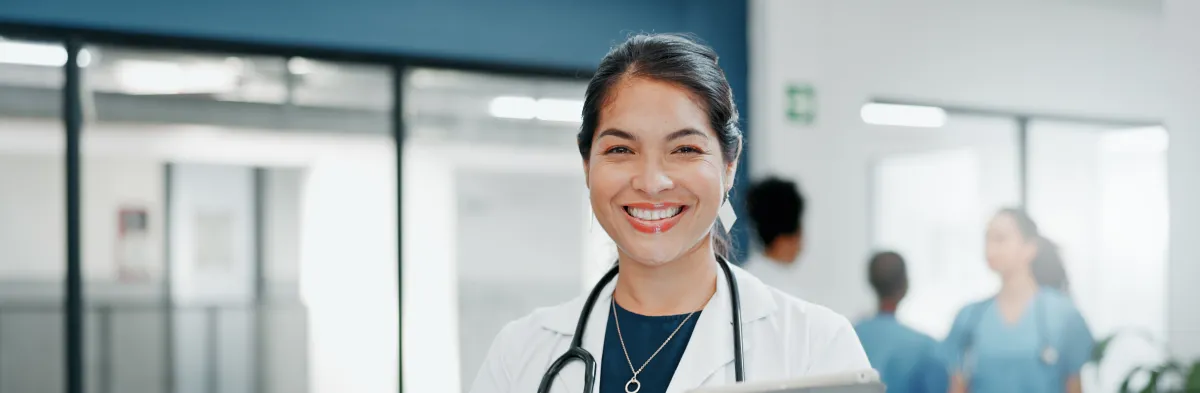 Smiling female nurse with a DNP wearing a white coat and stethoscope stands in a medical setting, holding a tablet, with healthcare professionals blurred in the background.