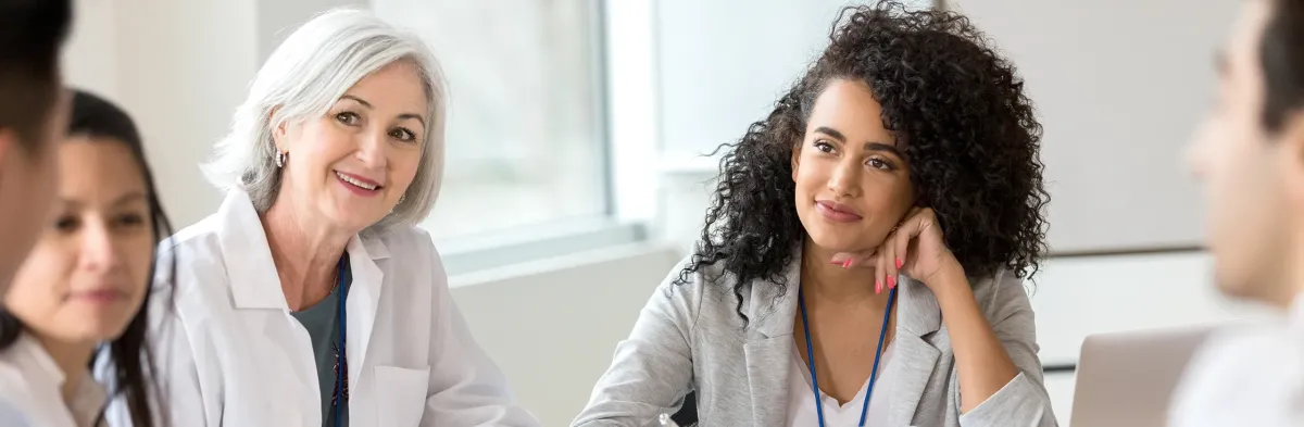 Two professional women with MSN degrees in nursing leadership and administration, one in a white lab coat and one in business attire, sit at a table engaged in discussion with colleagues, smiling and listening attentively.