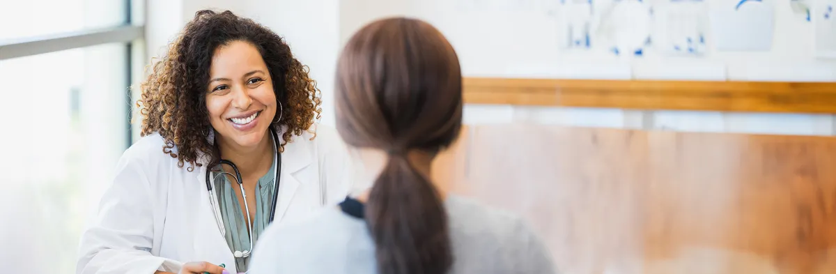 Smiling female nurse administrator in a white coat with a stethoscope talks with a staff member seated across from her in a bright medical office.
