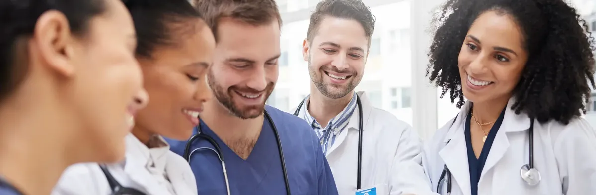 A group of smiling healthcare professionals, including doctors and nurses in white coats and scrubs with stethoscopes, gather together looking at a shared document or tablet.