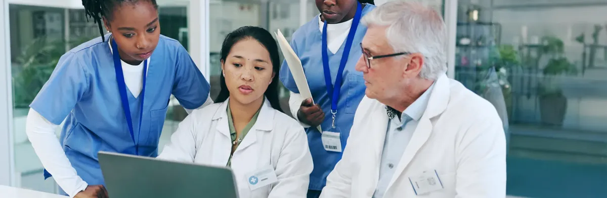 Nursing professionals in lab coats and scrubs gather around a laptop, reviewing information together, with one person holding a folder.