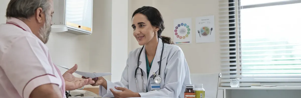 A female nurse who went from RN to DNP in a white coat and stethoscope smiles warmly while handing a paper to an older male patient during a consultation in a medical office