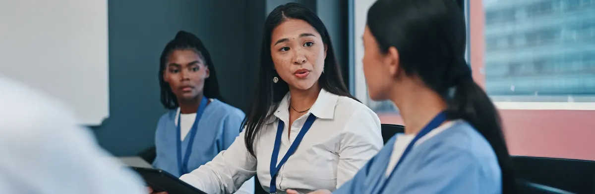 A woman who went from BSN to nursing administration in professional attire speaks to two nurses in blue scrubs during a team meeting, with another nurse listening in the background.