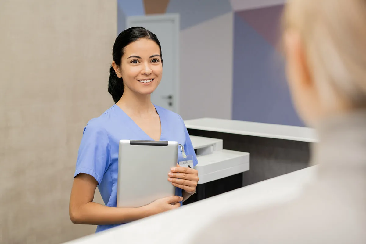 Medical assistant at front desk holding tablet smiling with patient
