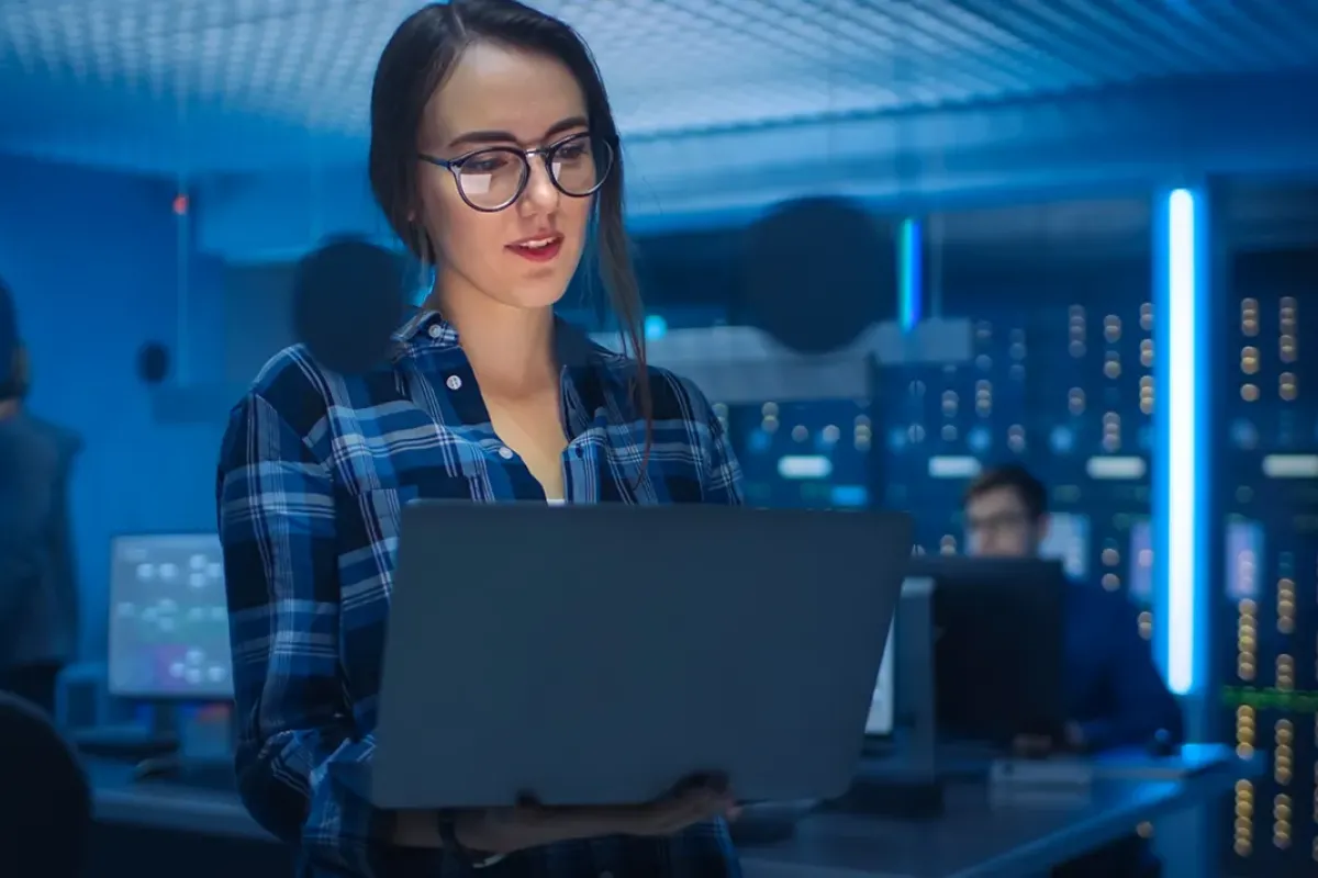 Cybersecurity analyst holding laptop in server room with coworkers in background