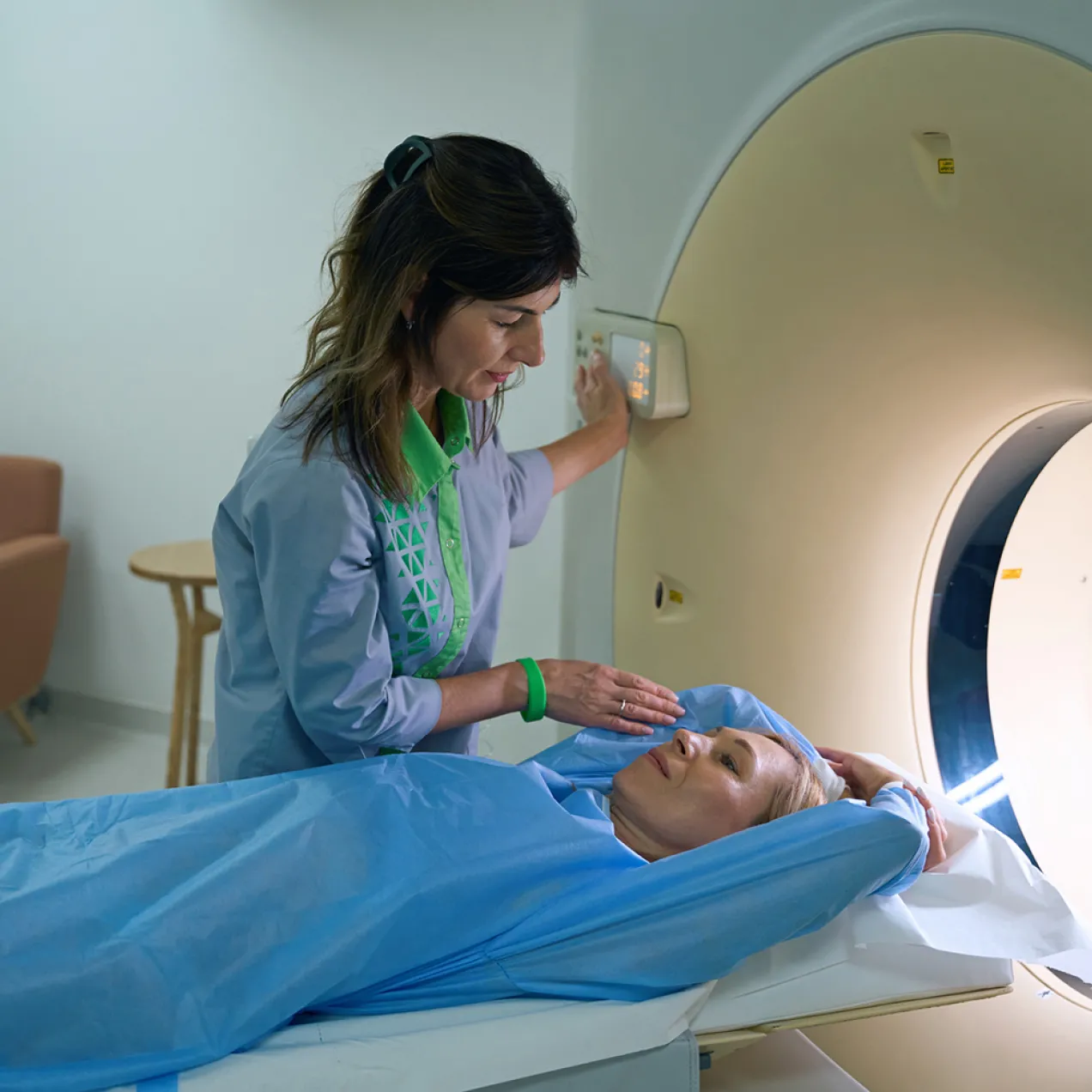 A radiologic technologist prepares a patient for a CT scan, adjusting the machine and providing reassurance as the patient lies on the exam table with arms raised above her head.