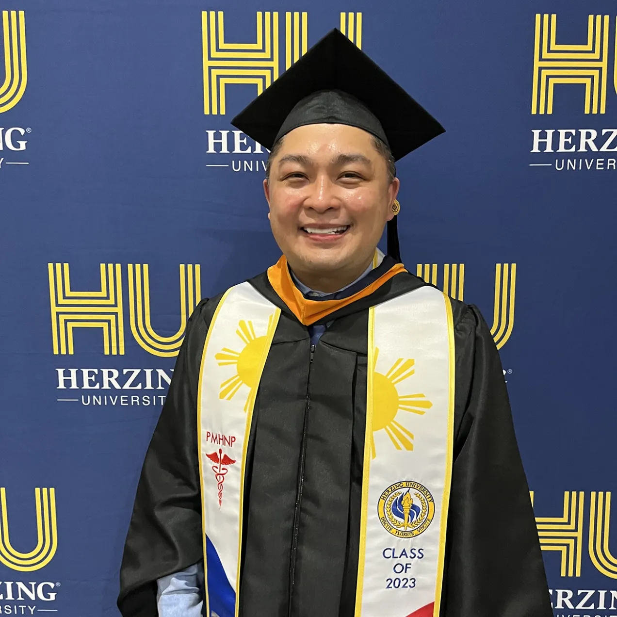 Graduate from Herzing University wearing a cap and gown with a Filipino flag stole, smiling and standing in front of a Herzing University banner. The stole features a PMHNP designation, indicating completion of the MSN-PMHNP program, Class of 2023.”