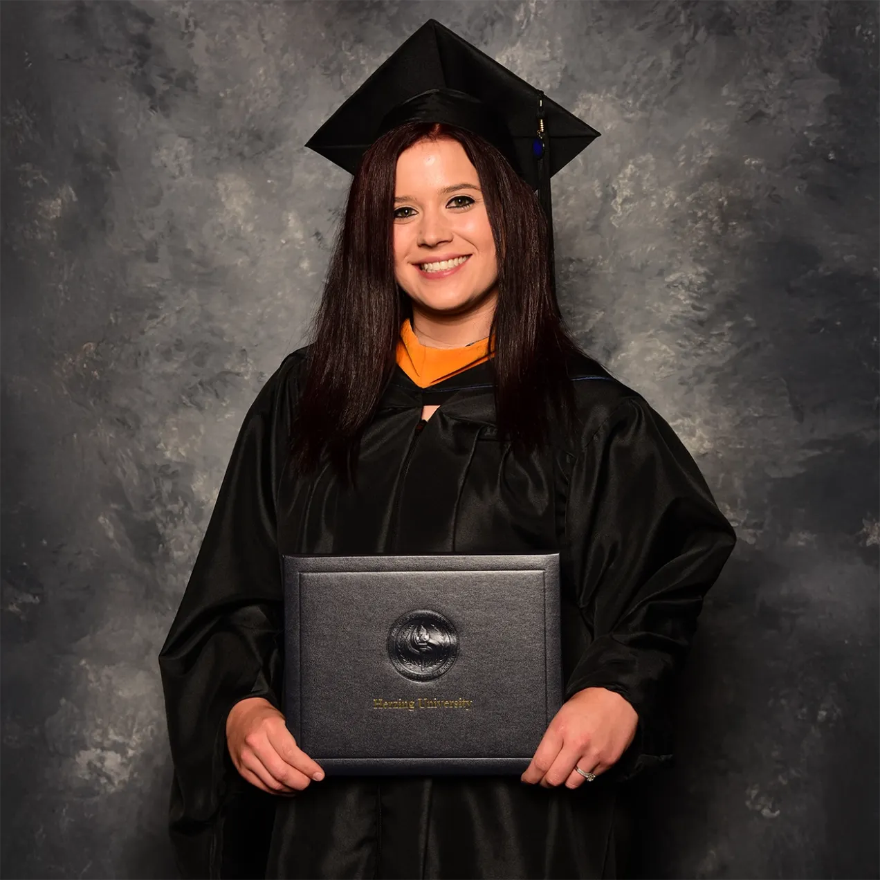 An appropriate alt tag for the image you uploaded could be: “Herzing University graduate holding a diploma in MSN-PMHNP program, wearing a graduation cap and gown, and smiling proudly.