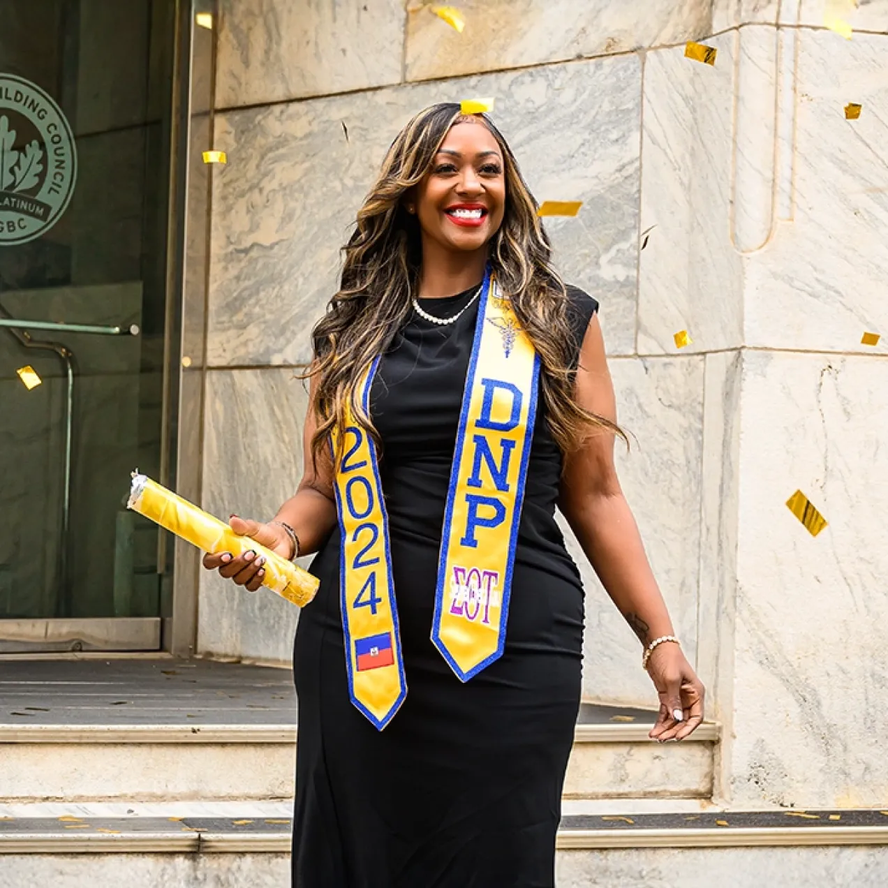 Graduating student celebrating with confetti, wearing a black dress and DNP 2024 sash, holding a diploma outside a building.