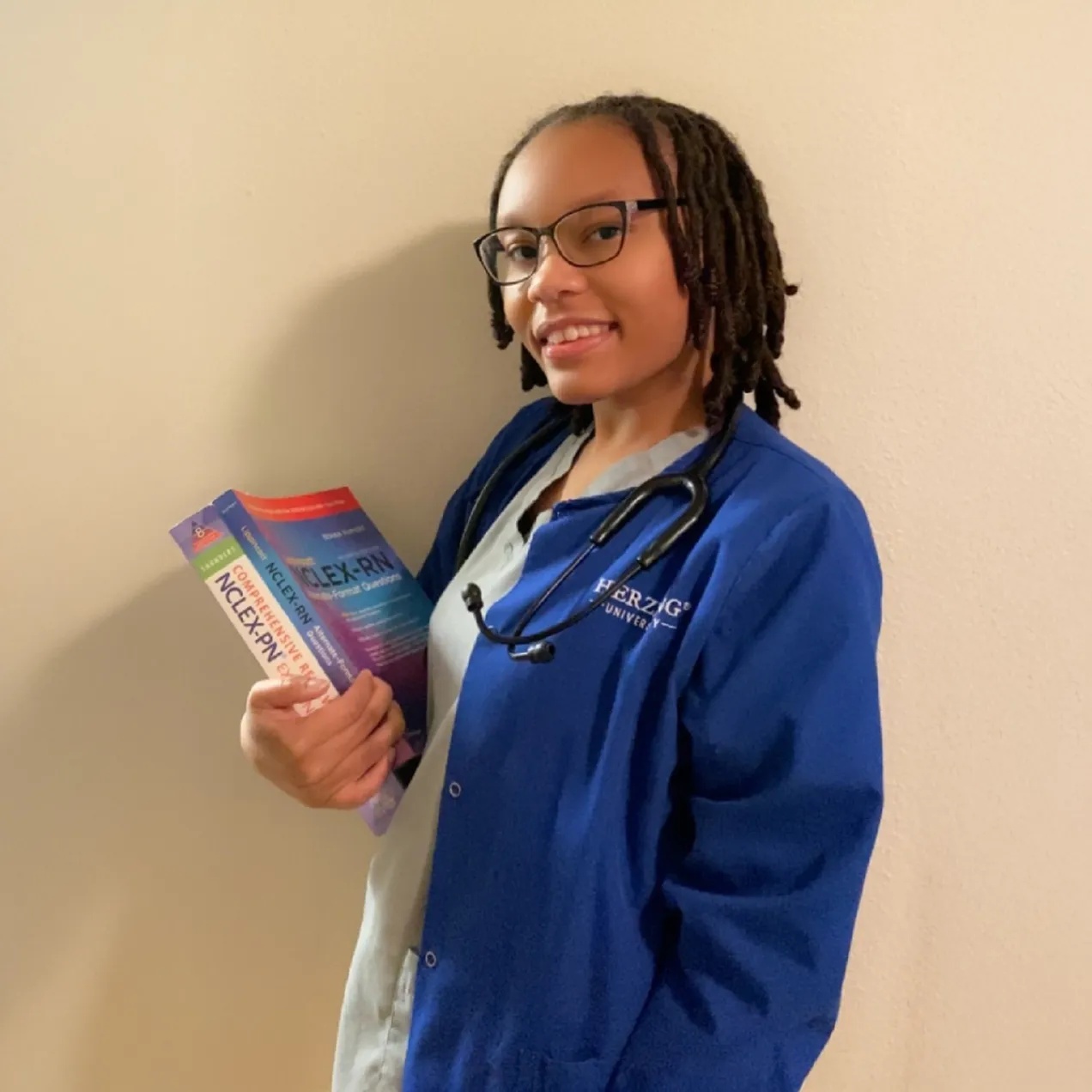 A Herzing University nursing student wearing a blue Herzing lab coat, stethoscope, and glasses, holding NCLEX exam preparation books while smiling confidently.