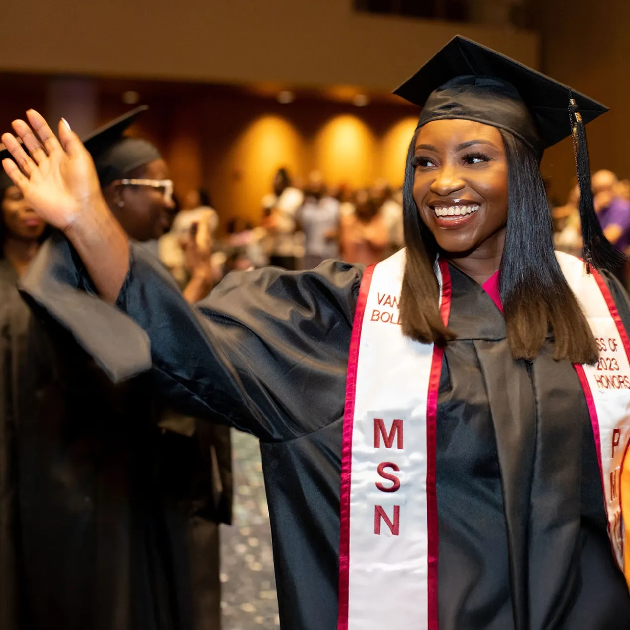 Graduate waving and smiling at a Herzing commencement ceremony, wearing an MSN sash, celebrating the completion of a Master of Science in Nursing degree.