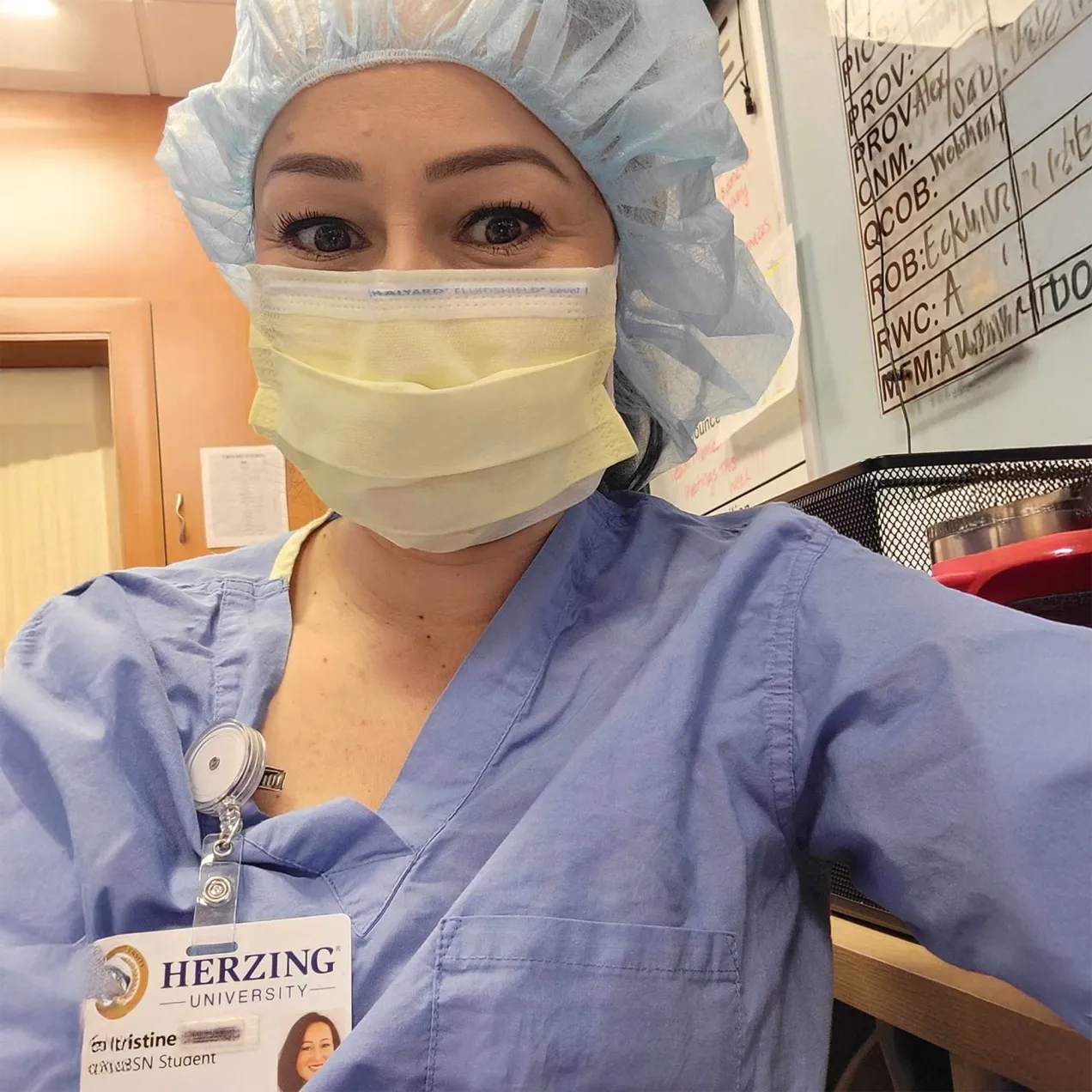 Surgical student in full PPE, wearing a Herzing University badge, smiling while taking a break during clinical training.