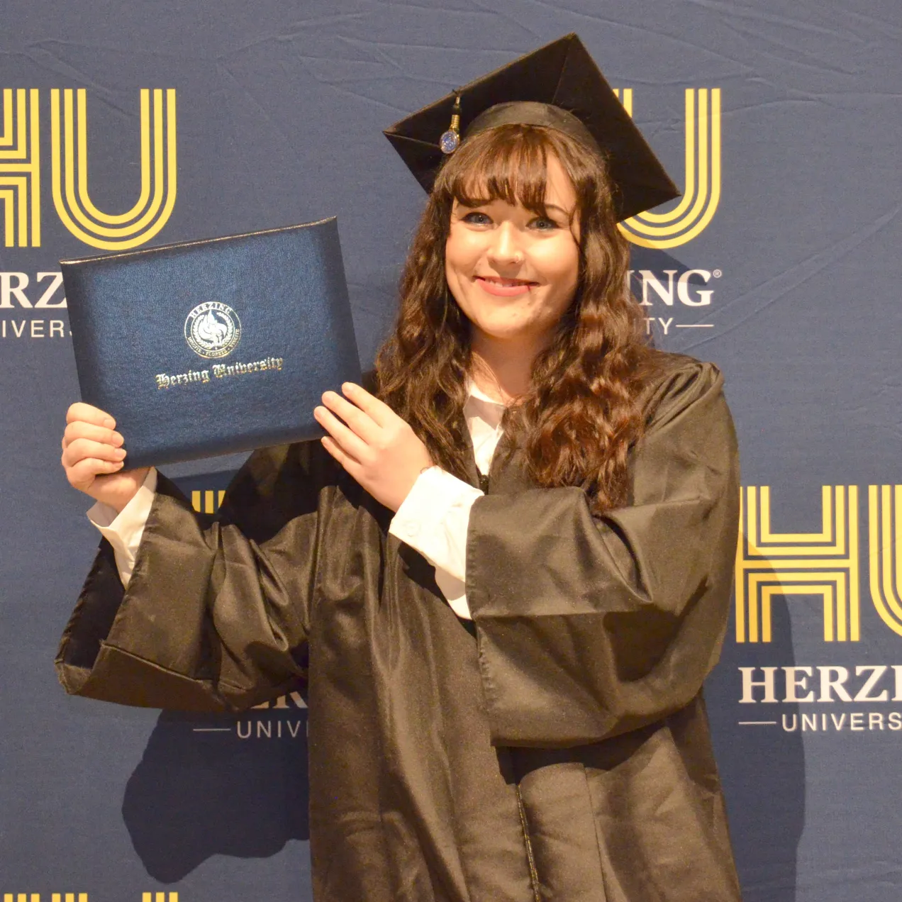Graduate smiling and holding diploma in front of Herzing University backdrop.