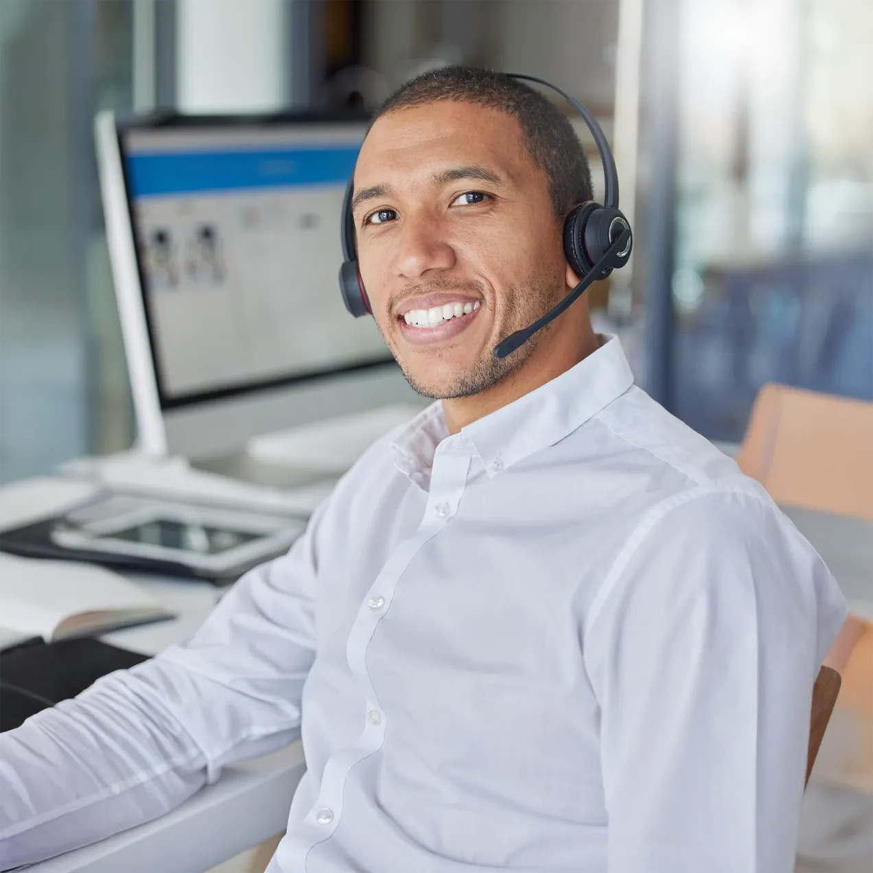 A Herzing University IT certificate student in a white shirt and headset smiles while providing technical support at a workstation with multiple screens.