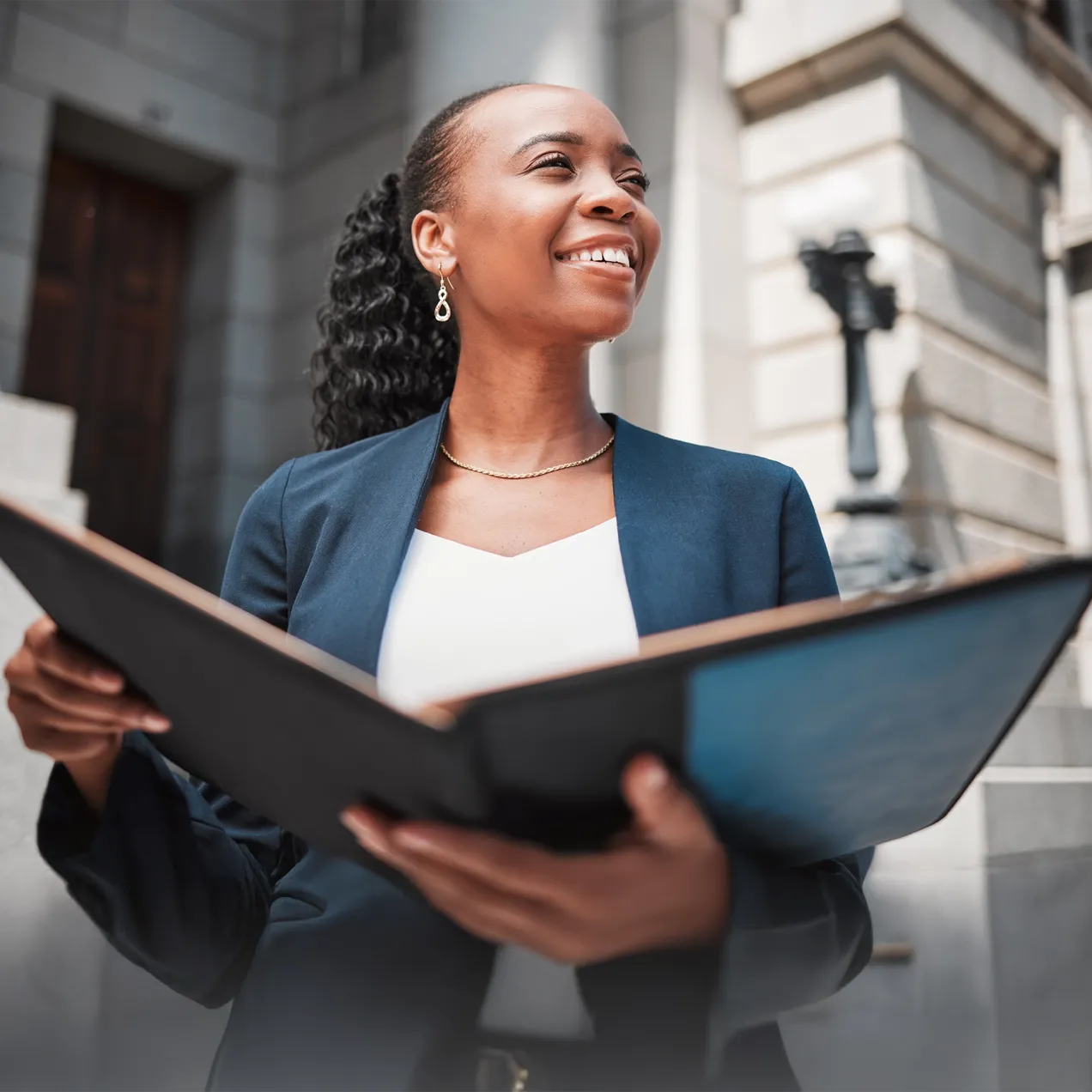 A confident woman in a navy blazer standing outside a courthouse, holding a portfolio, symbolizing career paths in law and legal studies at Herzing University.