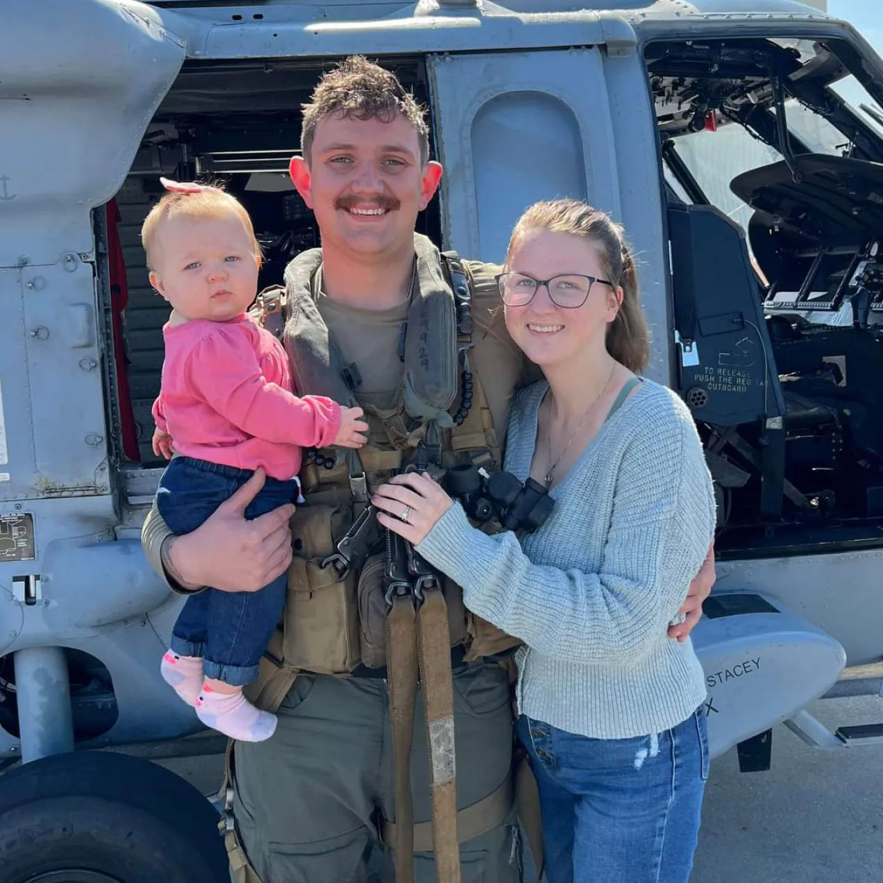 Military service member in tactical gear posing with family in front of a helicopter, representing service and family support.