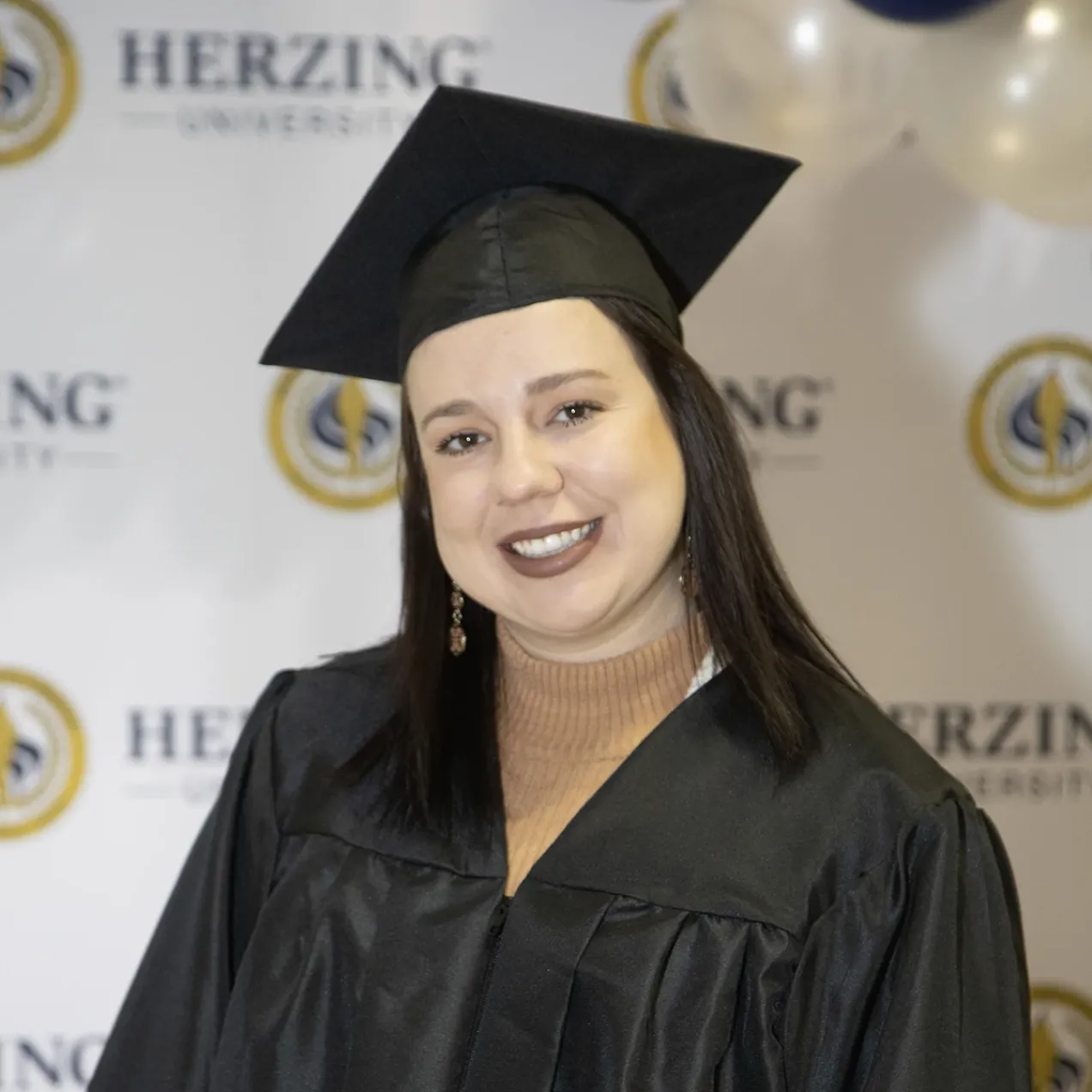 Herzing University graduate smiling in cap and gown at commencement, standing in front of official university backdrop.