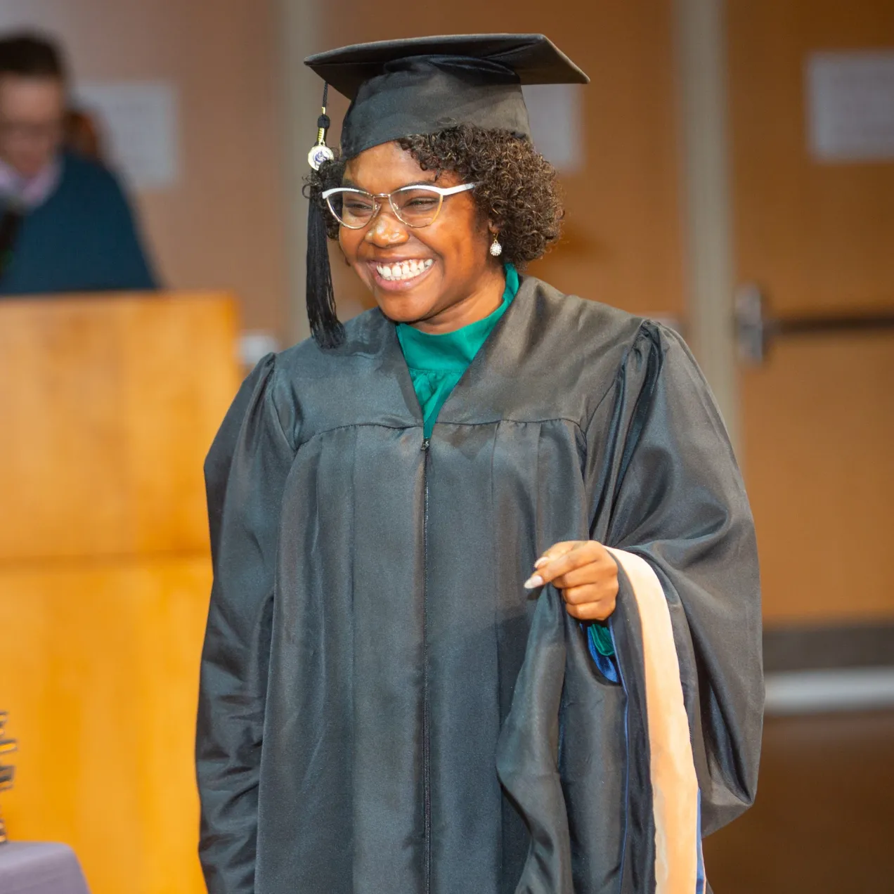Smiling Herzing University graduate in cap and gown holding academic hood during commencement ceremony.