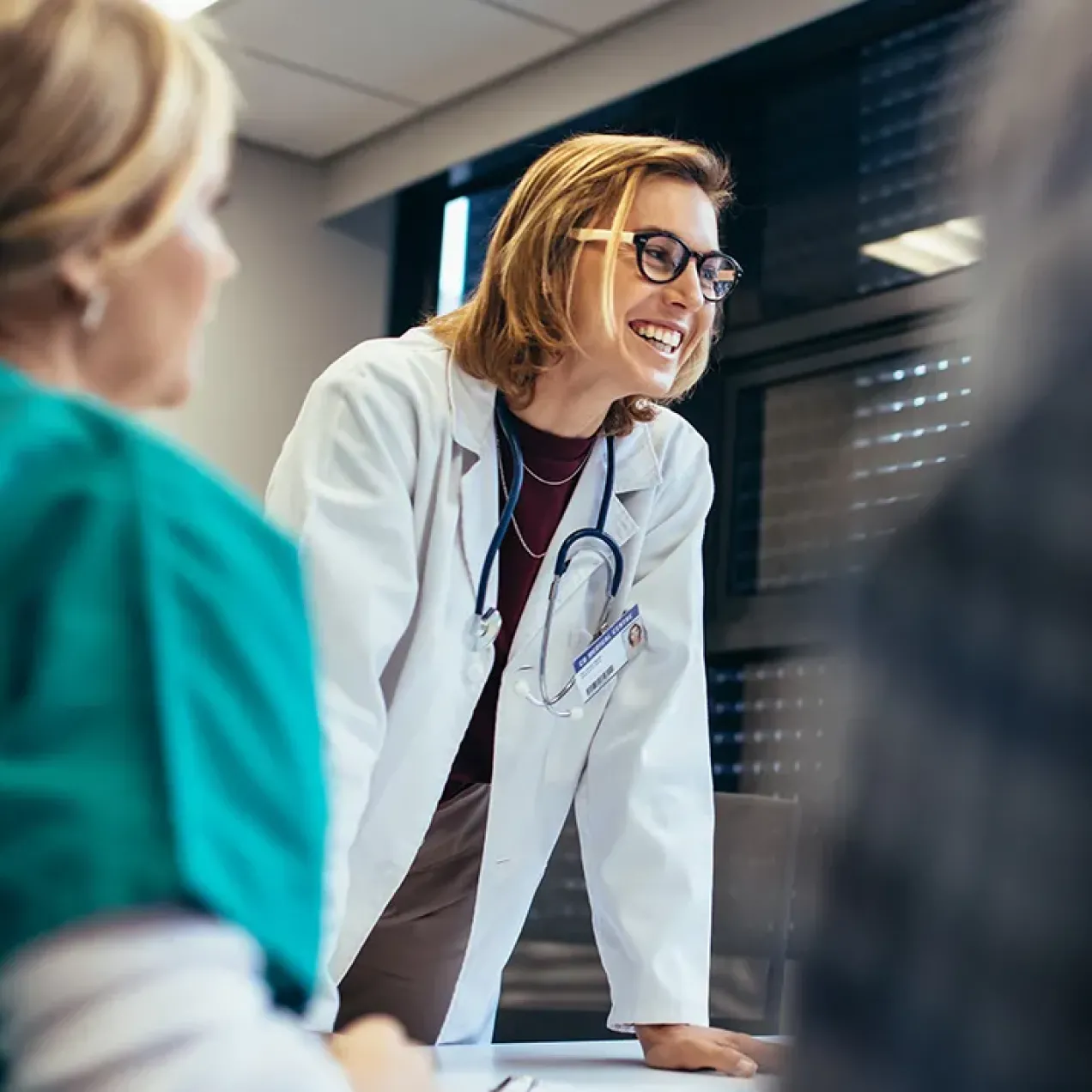 Smiling female healthcare educator in a white coat with a stethoscope leaning on a table, engaging with nursing students during a classroom discussion