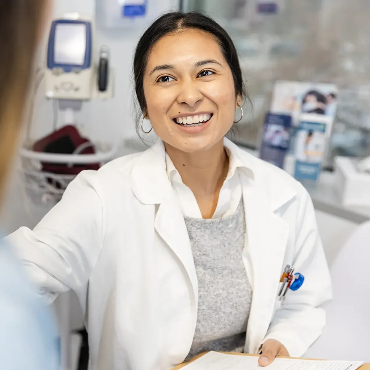 Smiling healthcare provider in a white coat speaking with a patient in an exam room while holding a clipboard