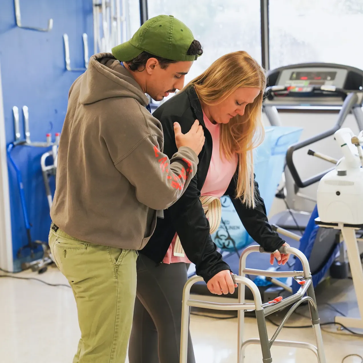Student assists a woman using a walker in a clinical training lab with exercise equipment in the background