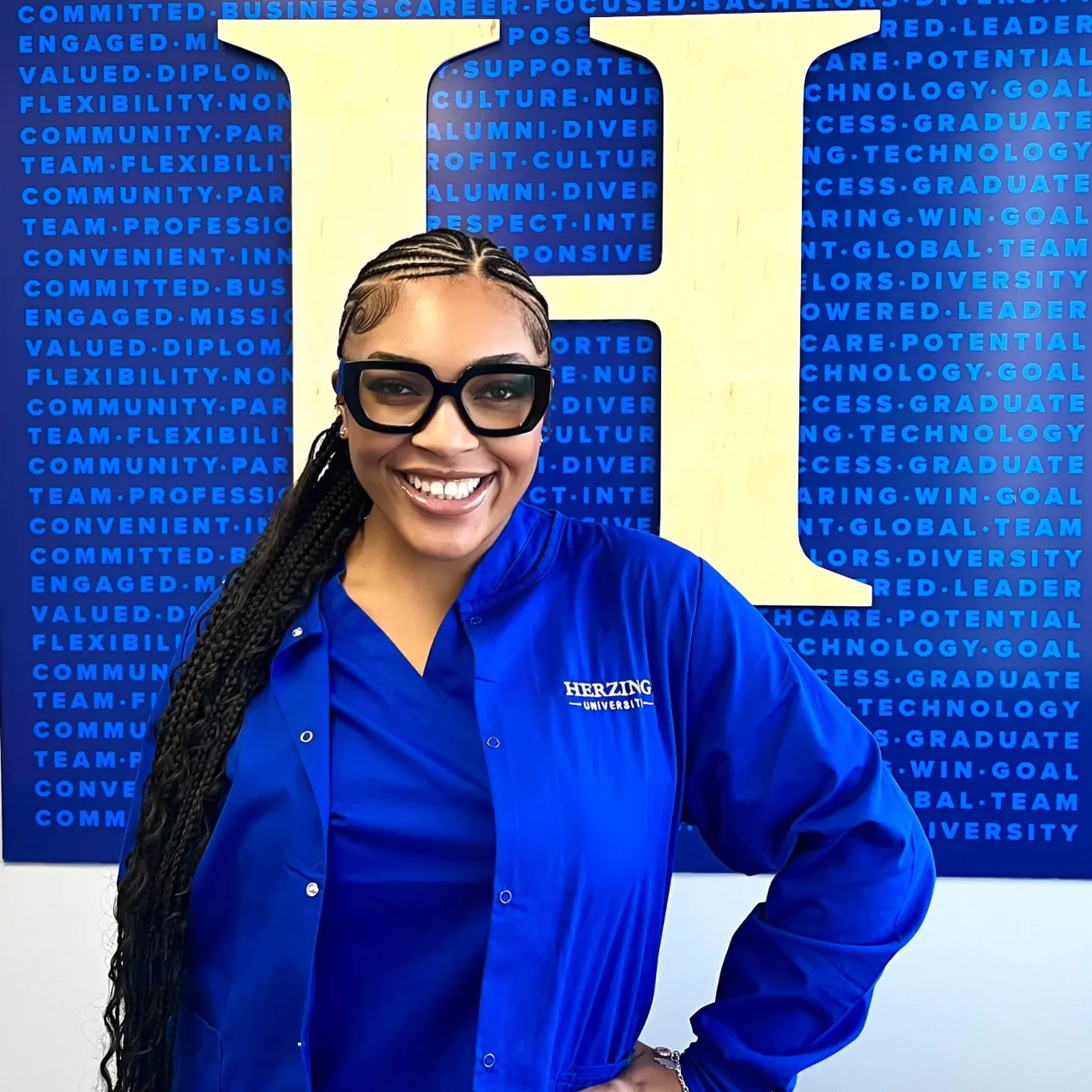 Herzing University Surgical Technology student in blue scrubs and oversized black glasses smiling in front of a blue and gold Herzing University wall display.