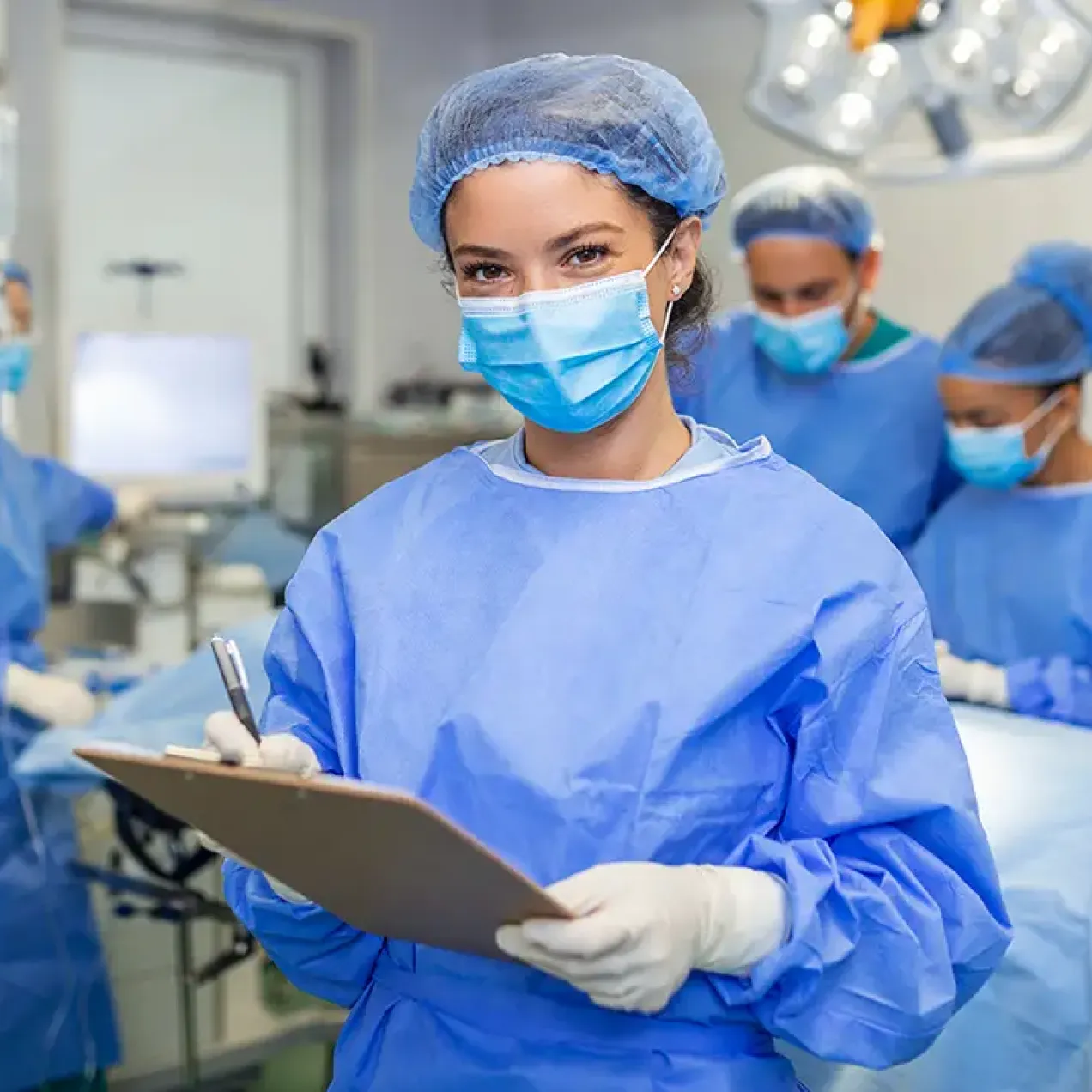 Surgical Technology student wearing protective surgical attire and holding a clipboard stands in an operating room while a clinical team prepares for a procedure in the background.