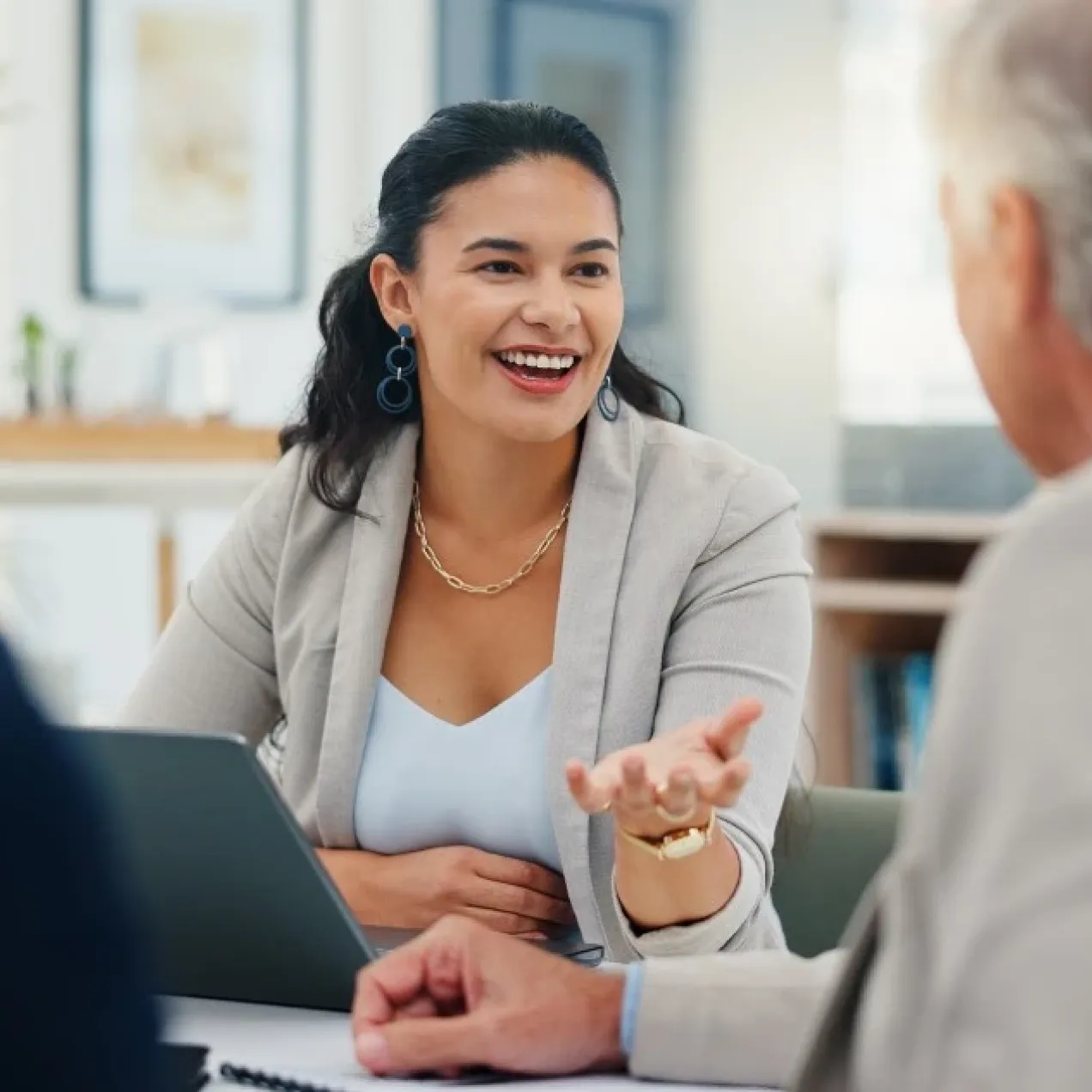 Smiling business professional speaks with two adults across a table during a meeting, gesturing while discussing information beside an open laptop in a bright office setting.