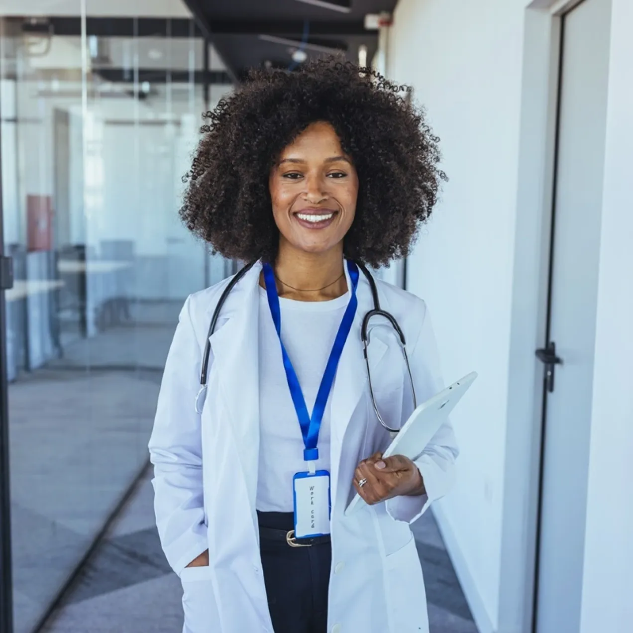 Smiling healthcare professional in a white coat with a stethoscope holds a tablet in a modern medical office.