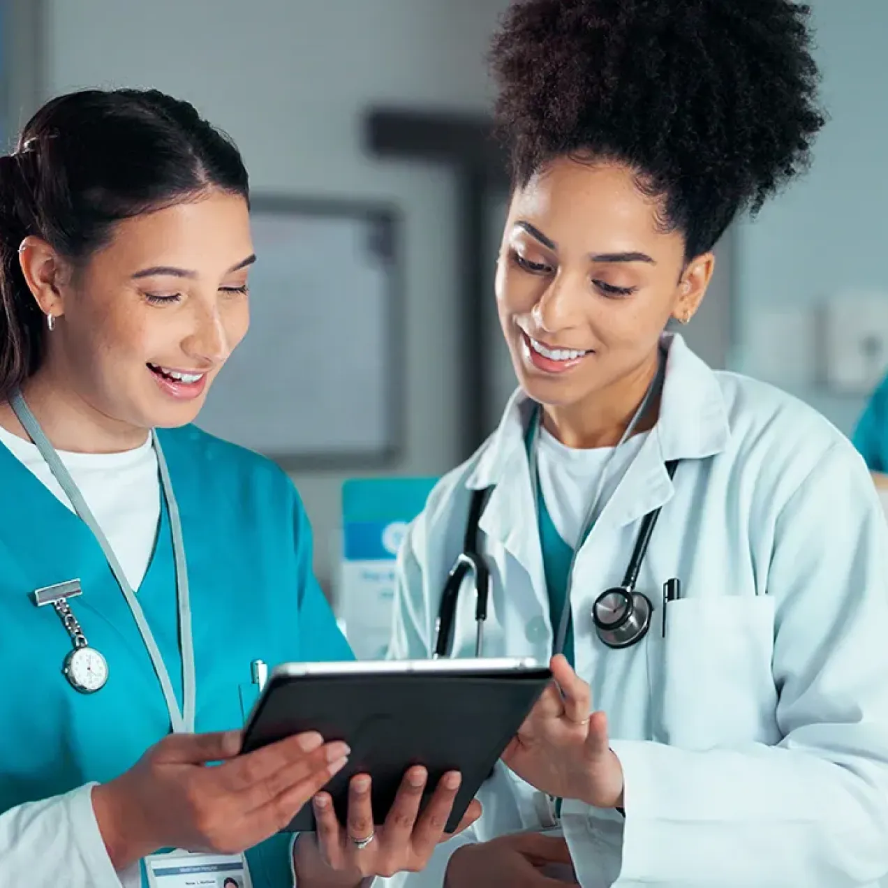 A nurse and clinician review information together on a tablet at a hospital workstation while another healthcare worker stands behind them.