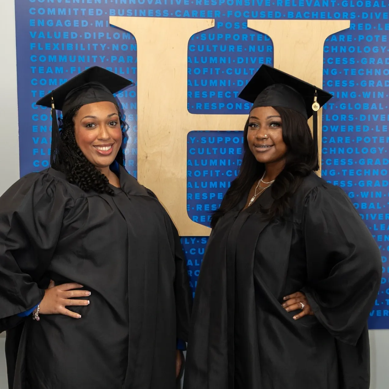 Two Herzing University graduates in cap and gown standing in front of Herzing logo at campus location