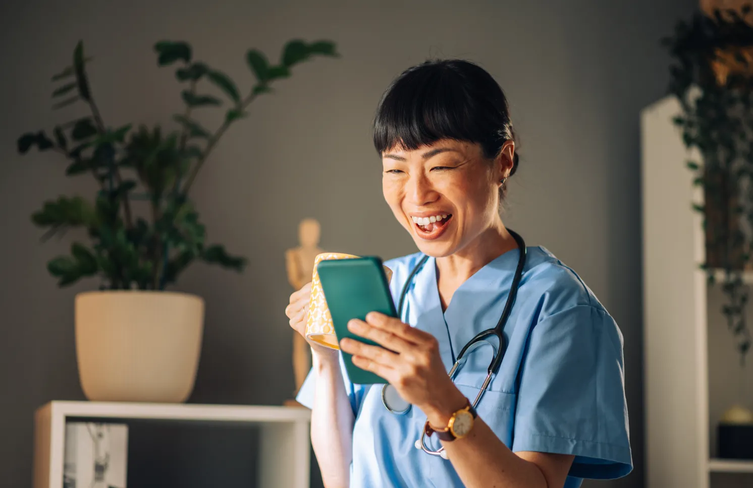 Asian female nurse in blue scrubs laughs heartily while looking at her mobile phone, holding a mug of coffee. The background features potted plants, bookshelves, and a small wooden figure.