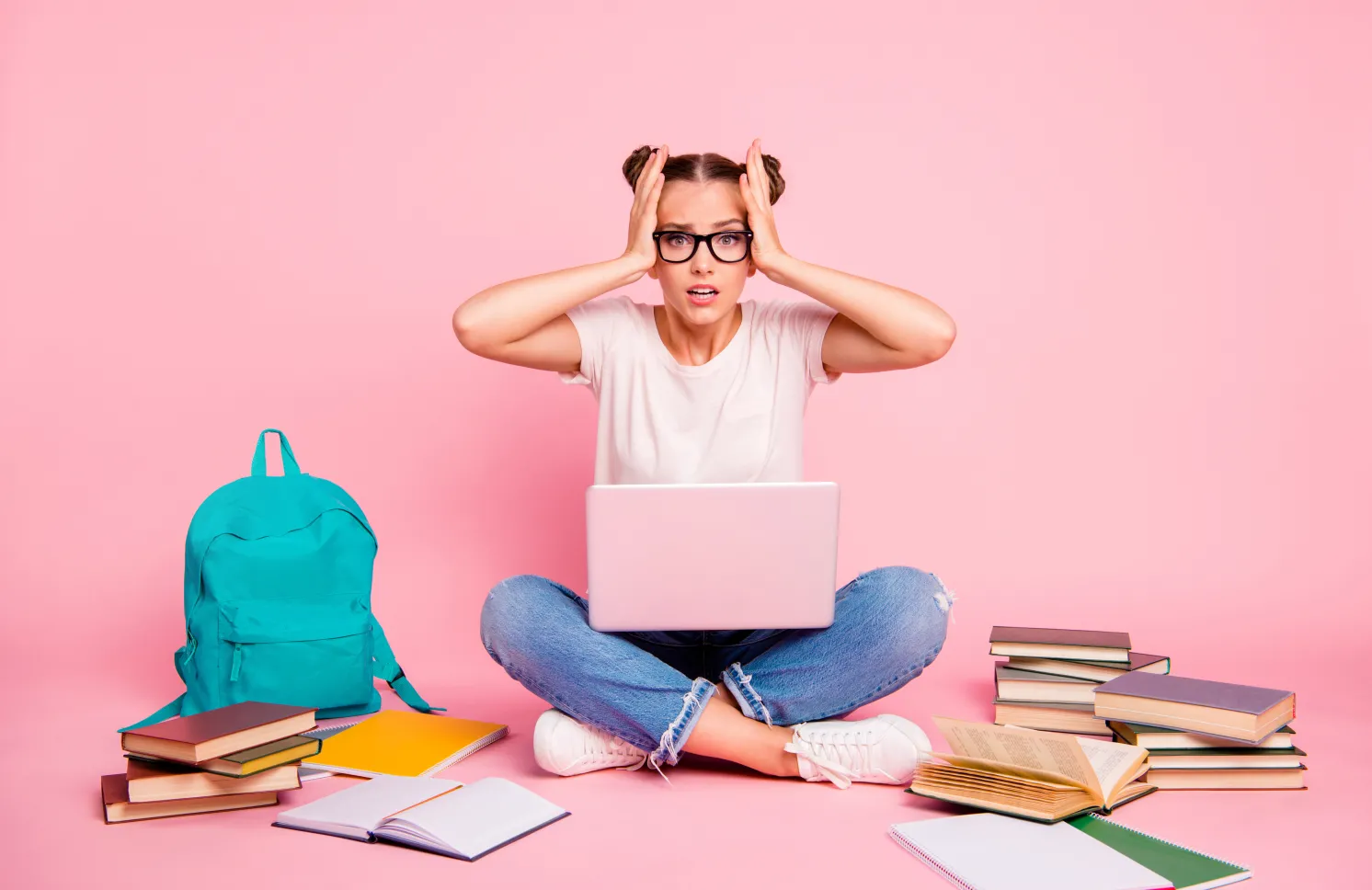 Full legs, body, size portrait of confused girl in white T-shirt and blue jeans sitting on floor with laptop on knees isolated on bright pink background.