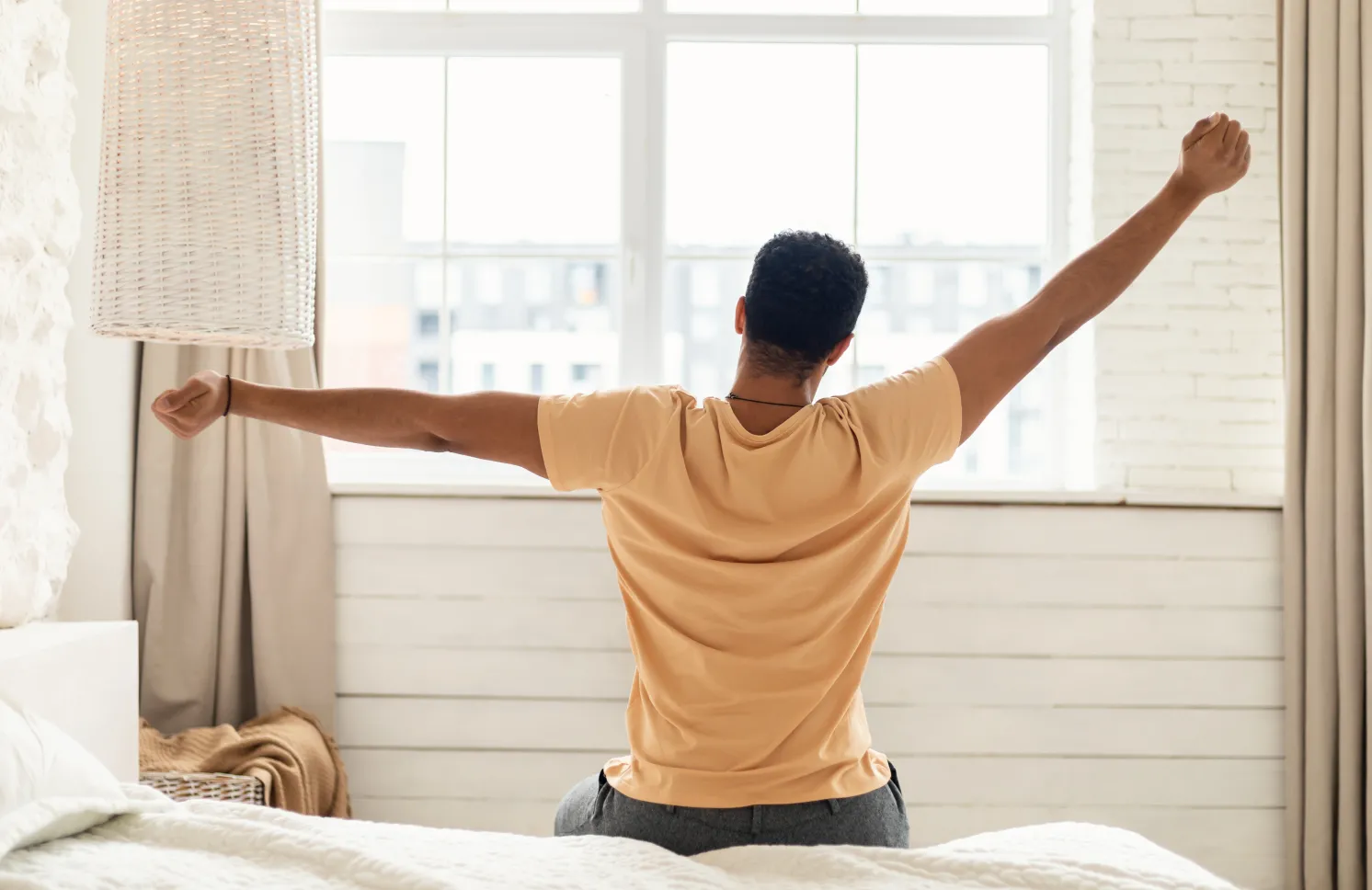 Arabic Man Sitting On Bed Back To Camera And Stretching Arms Awaking After Good Sleep In Modern Bedroom At Home, Looking Out Of Window. Comfort And Coziness. Good Morning Concept