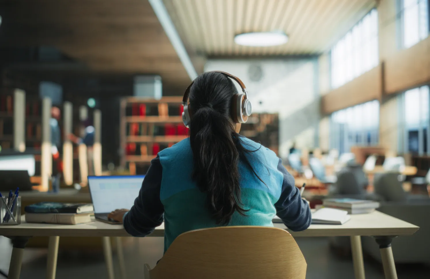 Female Student Wearing Headphones while Working on University Homework in a Public Library. Woman Sitting Behind a Desk, Using Laptop Computer and Writing Down Notes in Notebook. Footage from the Back