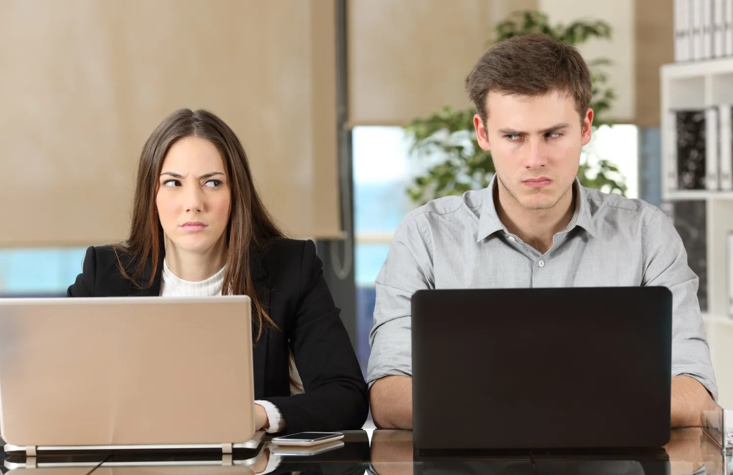 Front view of two angry businesspeople using computers disputing at workplace and looking sideways each other with anger