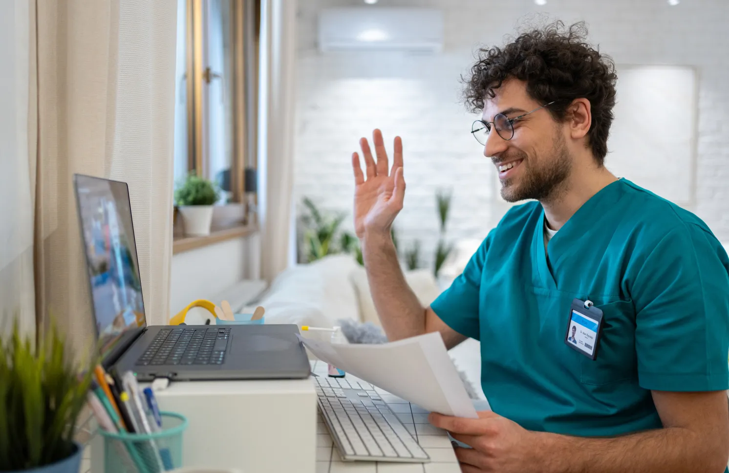 A young Caucasian male nurse in scrubs, having a video call while working from his home