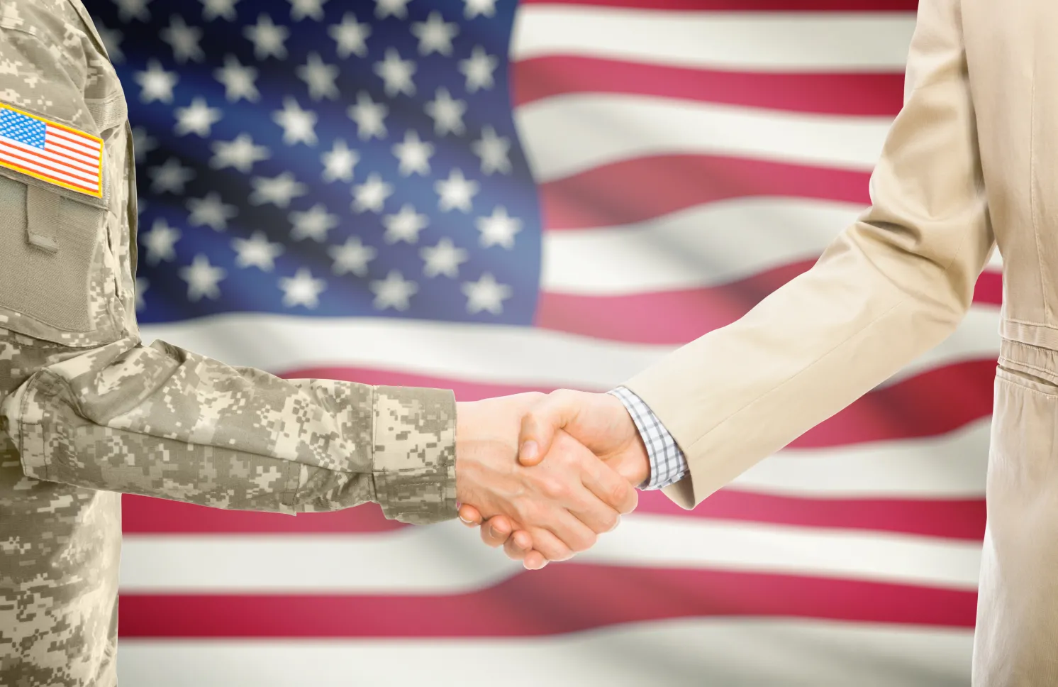 American soldier in uniform and civil man in suit shaking hands with national flag on background - United States