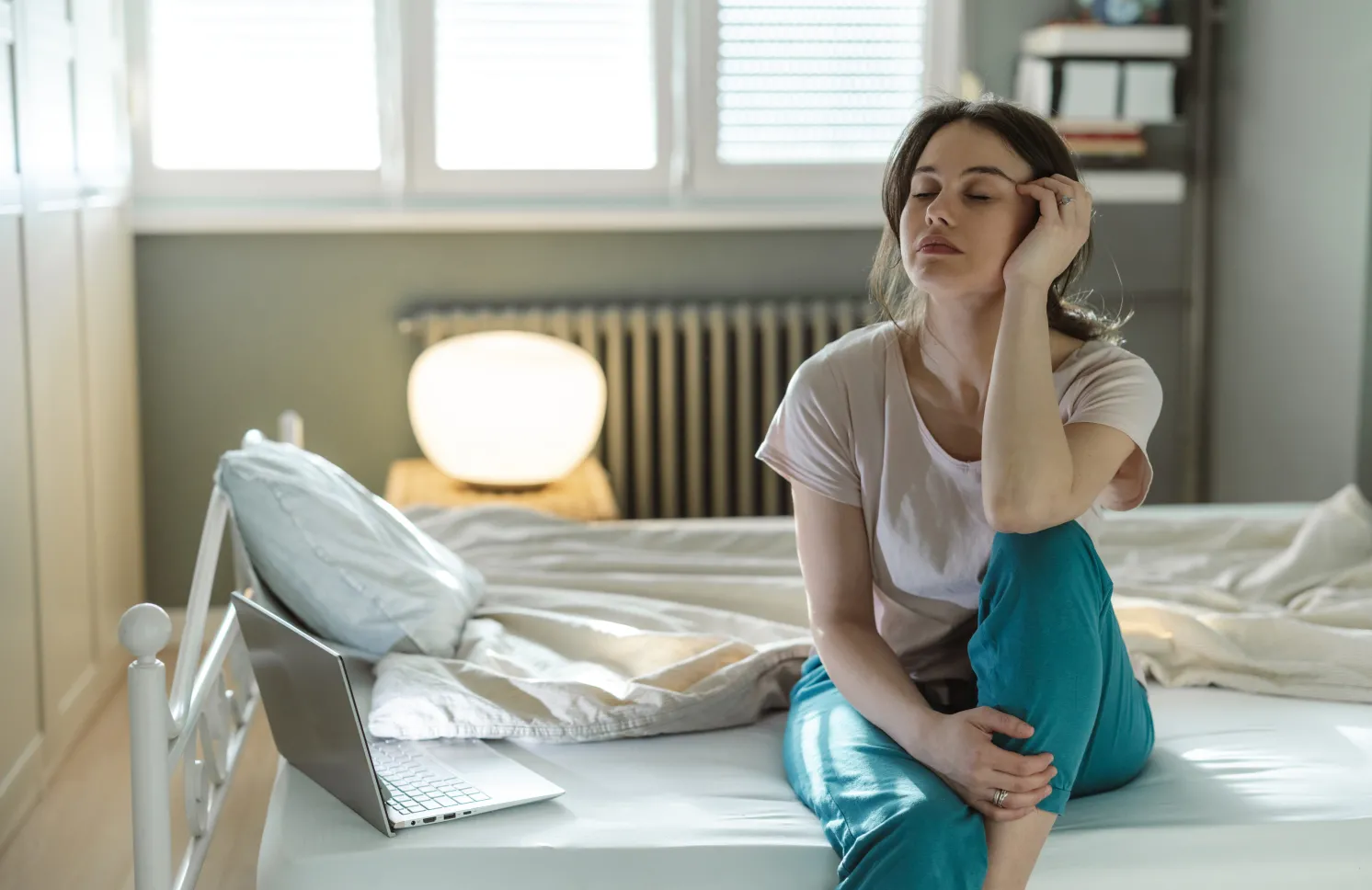 A young woman sitting on her bed in a sunny bedroom, appearing deep in thought and showing signs of tiredness. The scene suggests contemplation, relaxation, or a morning routine in a cozy setting.