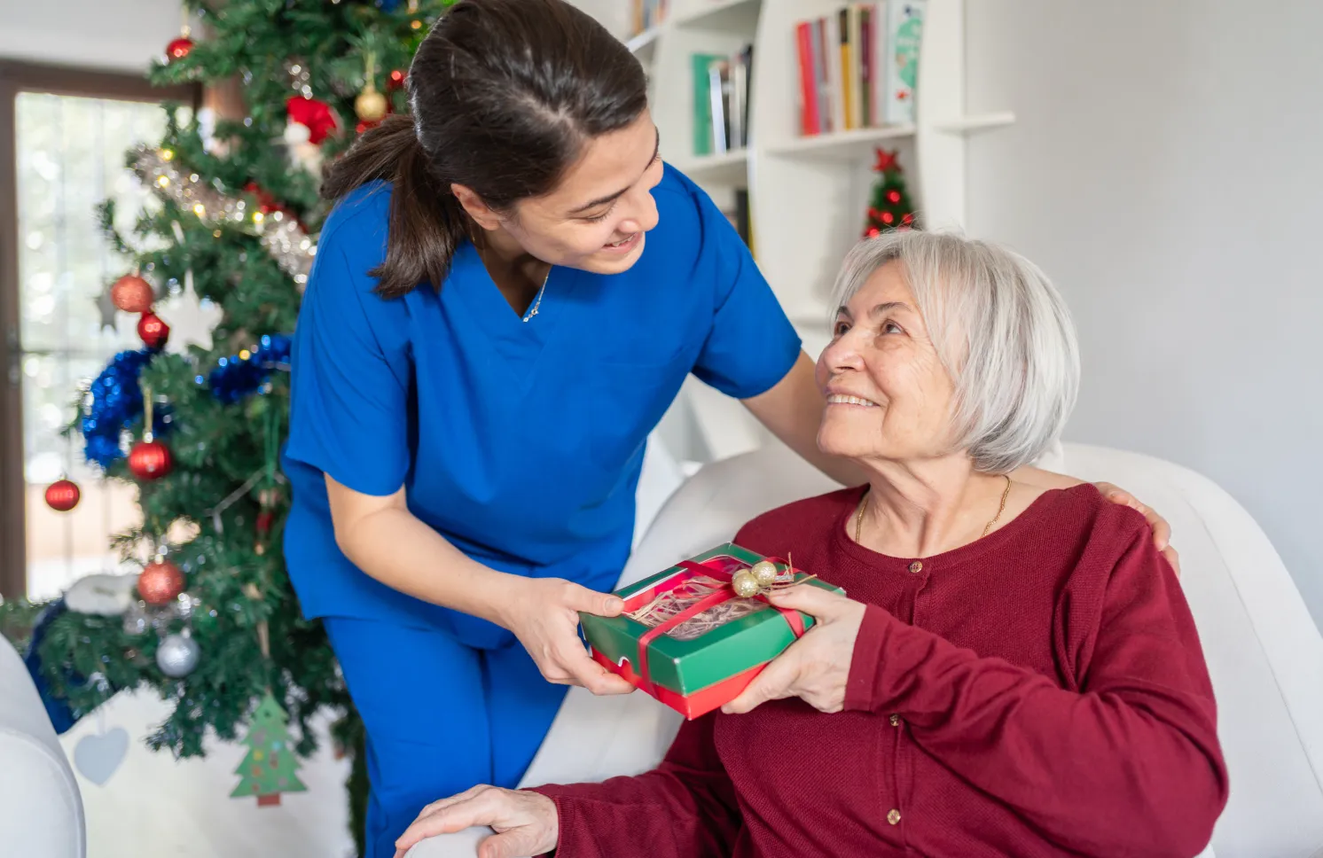 Home Caregiver Hugging And Giving Presents Senior Woman For Christmas At Nursing Home