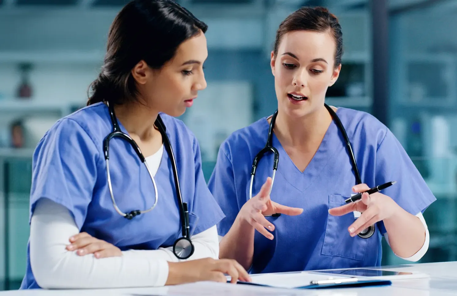 Two nurses in blue scrubs discuss treatment notes at a desk with a tablet and clipboard.