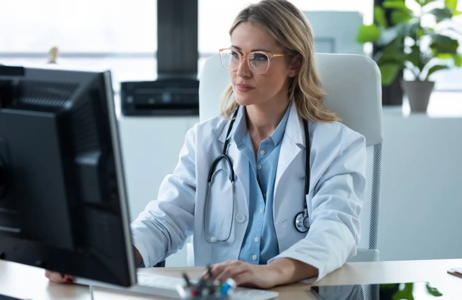 A nurse practitioner wearing a white lab coat and stethoscope sits at a desk using a computer in a bright medical office.