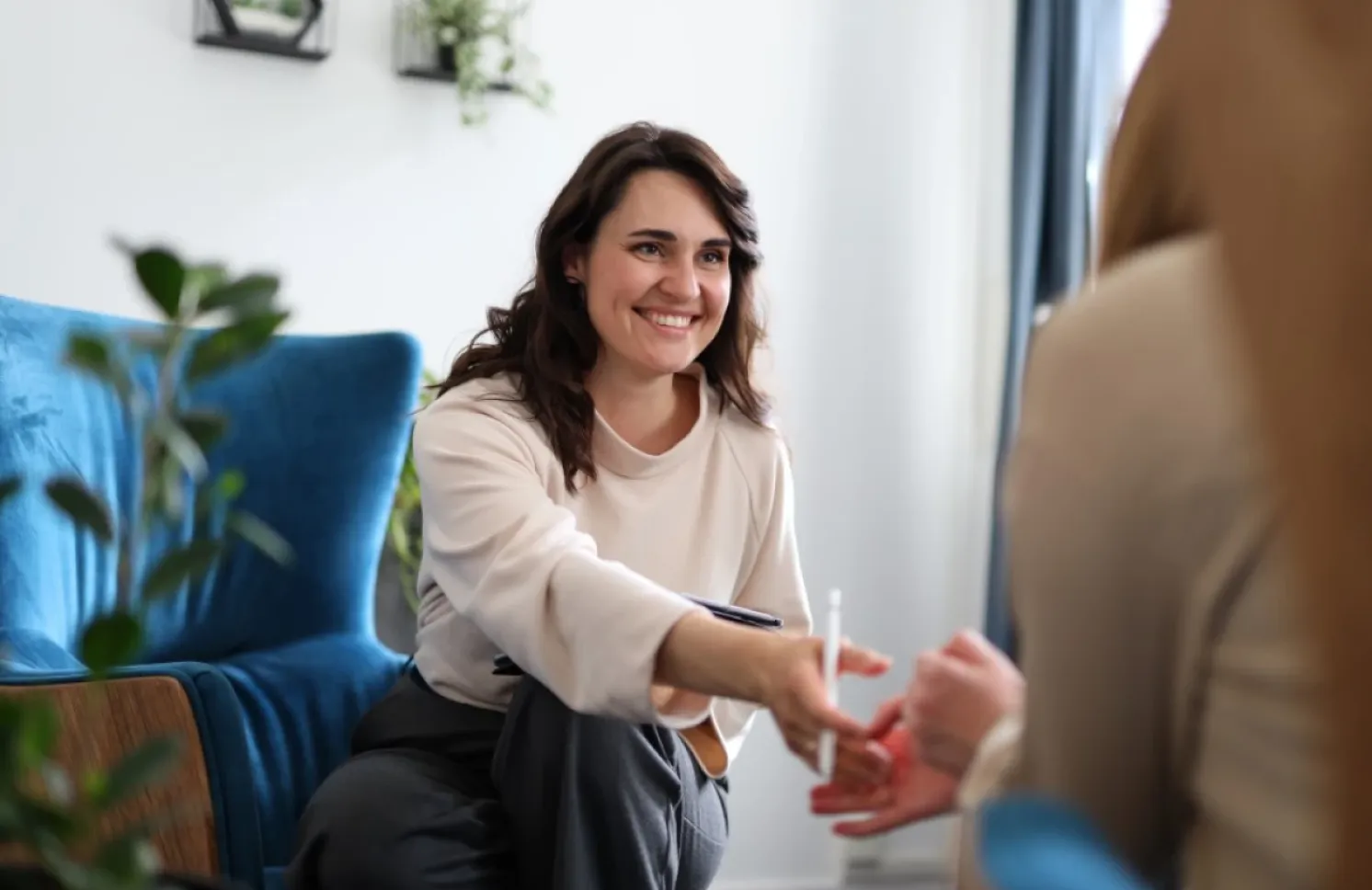 A smiling professional sits in a comfortable office, shaking hands with a client seated across from her. The room features a blue armchair, houseplants, and soft natural light, creating a welcoming and supportive environment.