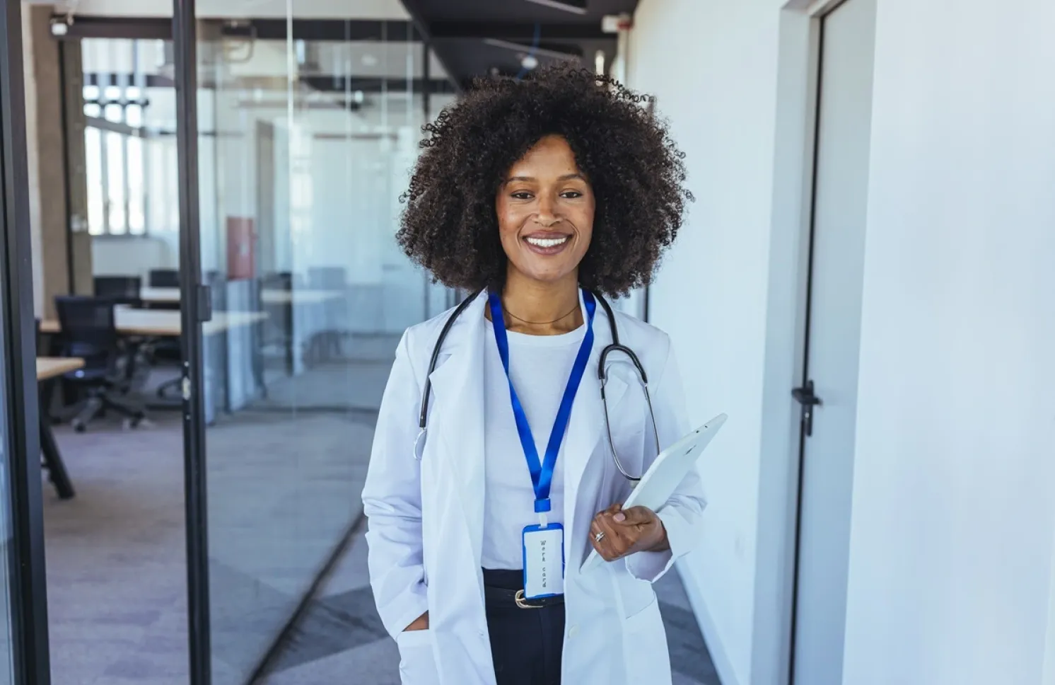 Smiling nurse practitioner in a white coat with a stethoscope holds a tablet in a modern medical office.
