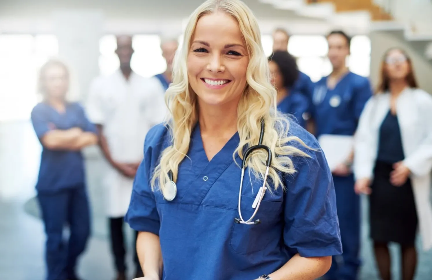 Smiling RN who is pursuing a BSN in blue scrubs with a stethoscope stands in the foreground while a multidisciplinary medical team gathers in the background of a healthcare facility.