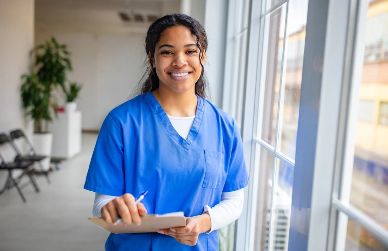 A smiling nurse in blue scrubs holds a clipboard and pen while standing beside large windows in a bright healthcare facility.