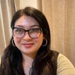 Latina woman in a yellow shirt poses in front of a curtain for a selfie